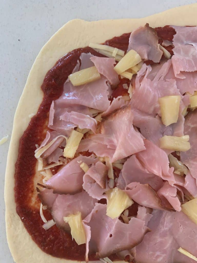 Un primer plano de la masa de pizza con pasta de tomate, jamón afeitado y piña en la parte superior. 