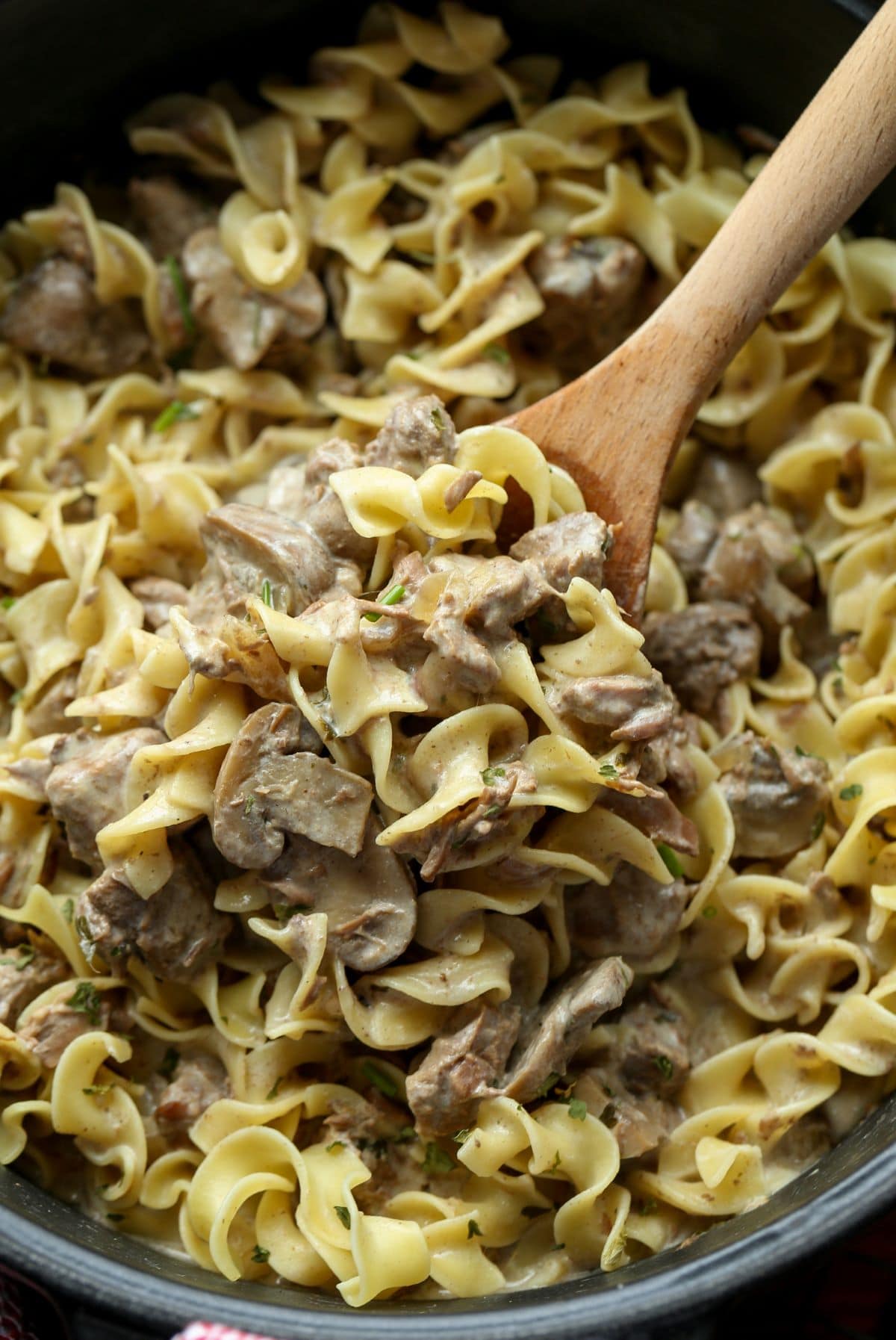 Una porción de stroganoff con fideos de huevo que se saca de una olla de cocción lenta con una cuchara de madera.