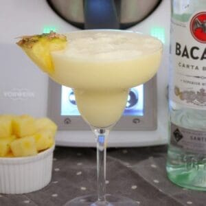 Un trozo de piña en el borde de un cóctel cremoso en una copa de cóctel con tallo, con una botella de Bacardi al lado.