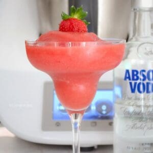 Una bebida de fresa fangosa servida en una copa de cóctel con tallo con una fresa encima y una botella de vodka cerca.