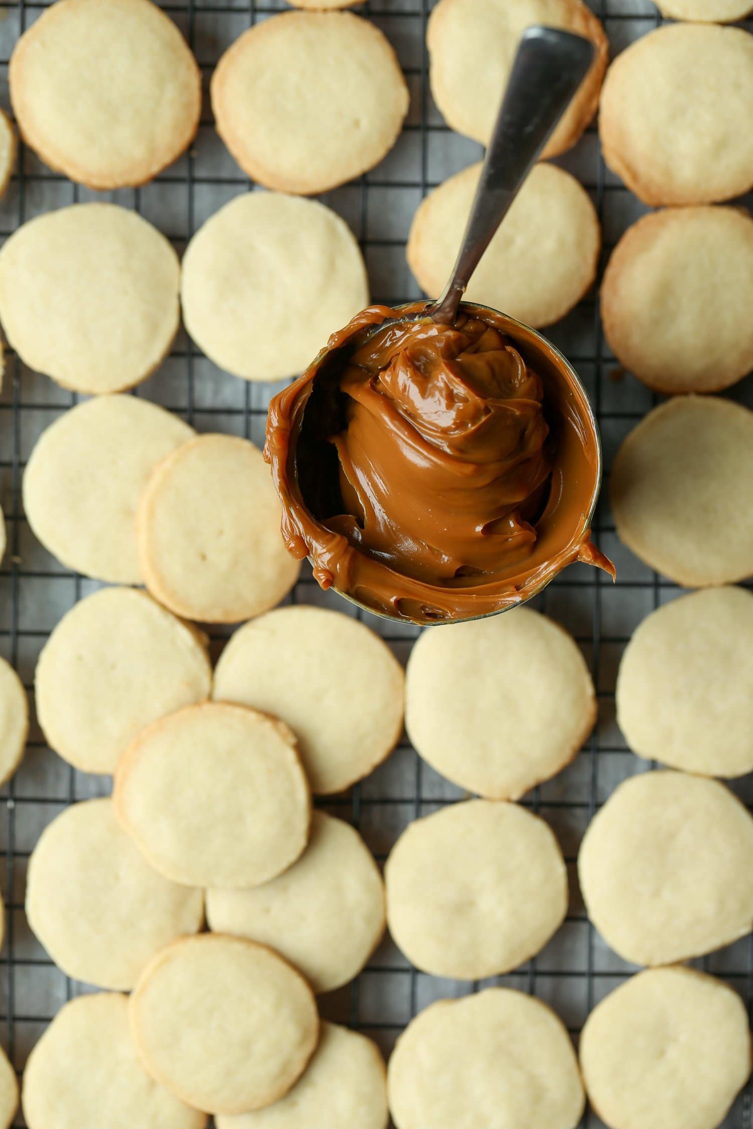 Una vista superior de un frasco de dulce de leche con una cuchara, rodeado de mitades de alfajores horneados.