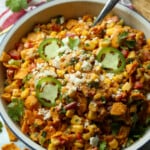 Ensalada picante de maíz Frito cubierta con queso cotija y jalapeños rebanados en una ensaladera blanca grande con una cuchara para servir.