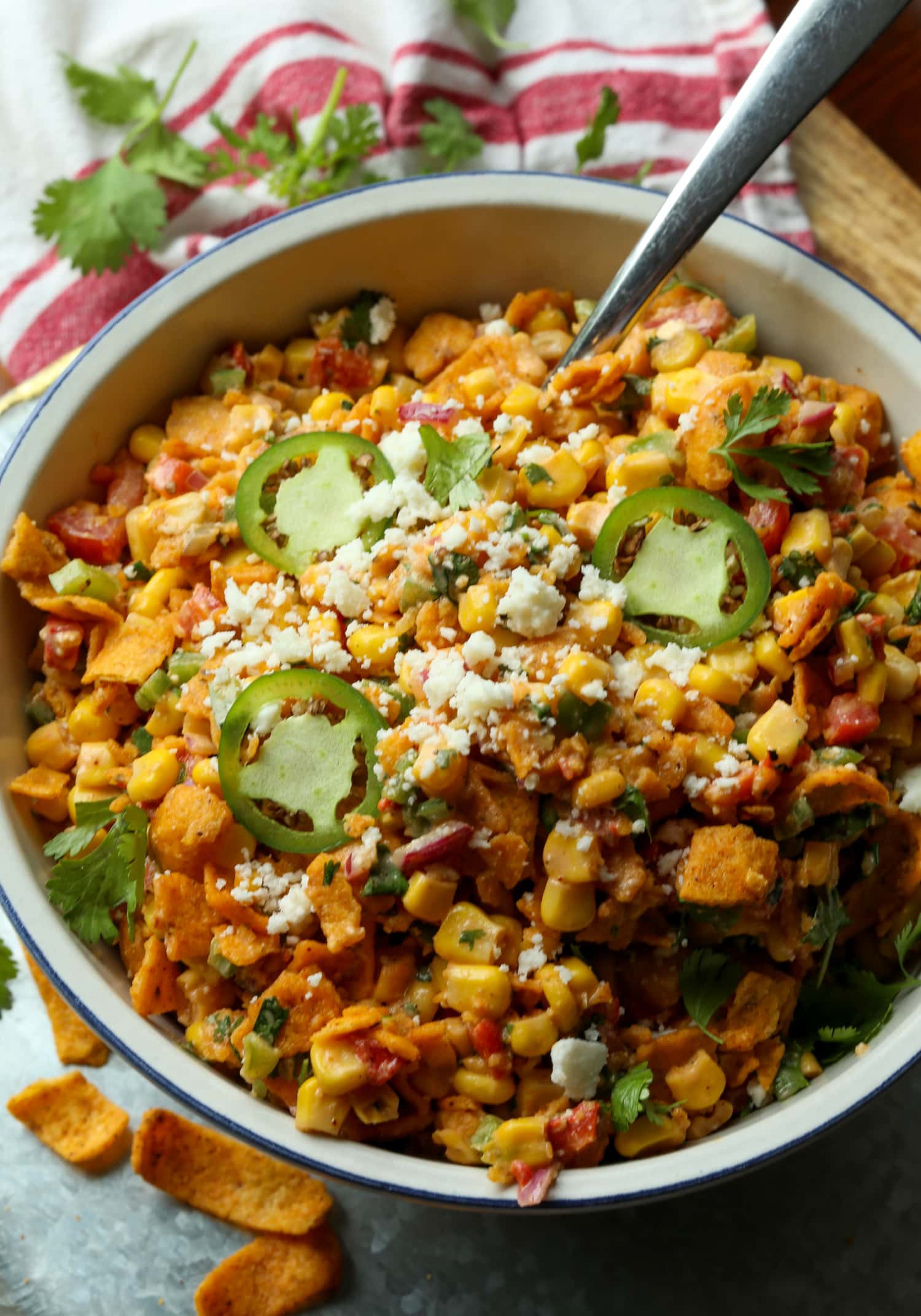 Ensalada picante de maíz Frito cubierta con queso cotija y jalapeños rebanados en una ensaladera blanca grande con una cuchara para servir.