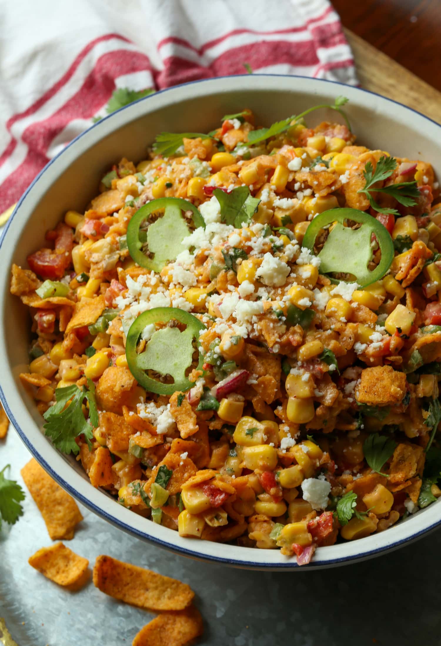 Spicy Frito Corn Salad es una receta de ensalada de verano fresca y sabrosa cargada de maíz dulce, verduras crujientes, chile picante y fritos crujientes. Ensalada picante de maíz Frito cubierta con queso cotija y jalapeños rebanados en una ensaladera blanca grande.