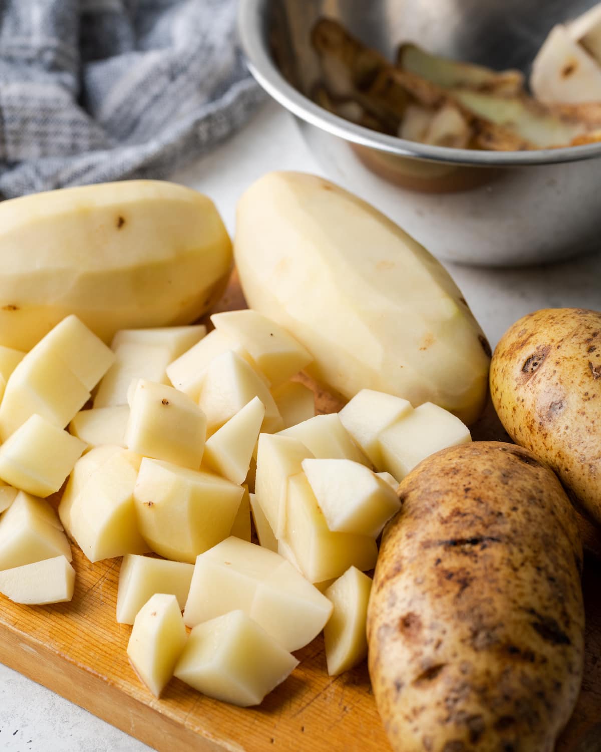 ¡Esta sencilla sopa instantánea de papas está lista en minutos! Una receta de sopa sedosa, cremosa y nutritiva cargada de queso derretido y verduras. Patatas picadas en una tabla de cortar junto a patatas enteras peladas y sin pelar.