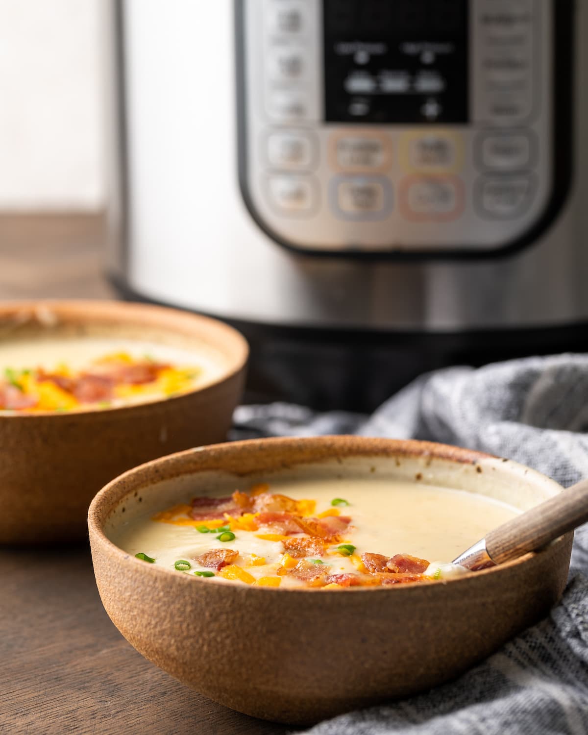 ¡Esta sencilla sopa instantánea de papas está lista en minutos! Una receta de sopa sedosa, cremosa y nutritiva cargada de queso derretido y verduras. Dos tazones de sopa de papa con guarnición de tocino, queso y cebollín con la olla instantánea al fondo.
