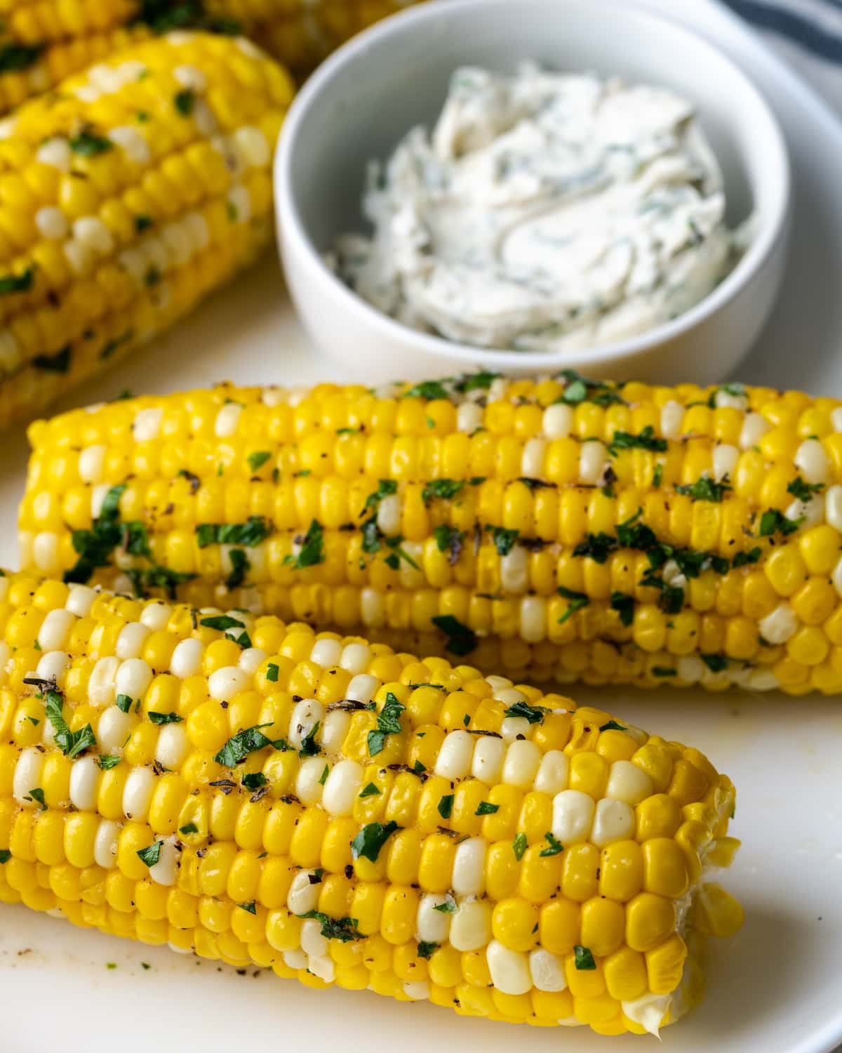 Instant Pot Corn on the Cob es, con mucho, la forma más fácil de disfrutar maíz fresco en minutos. Una guarnición fresca de verano cubierta con mantequilla de hierbas casera. Maíz en mazorca cubierto con mantequilla y hierbas, junto a un plato de mantequilla de hierbas.