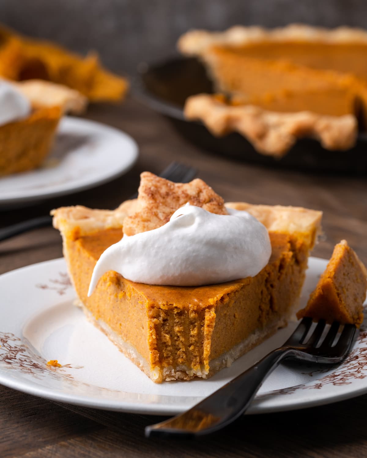 Un trozo de pastel de calabaza en un plato cubierto con crema batida y una galleta de azúcar, al que le falta un tenedor lleno de pastel.