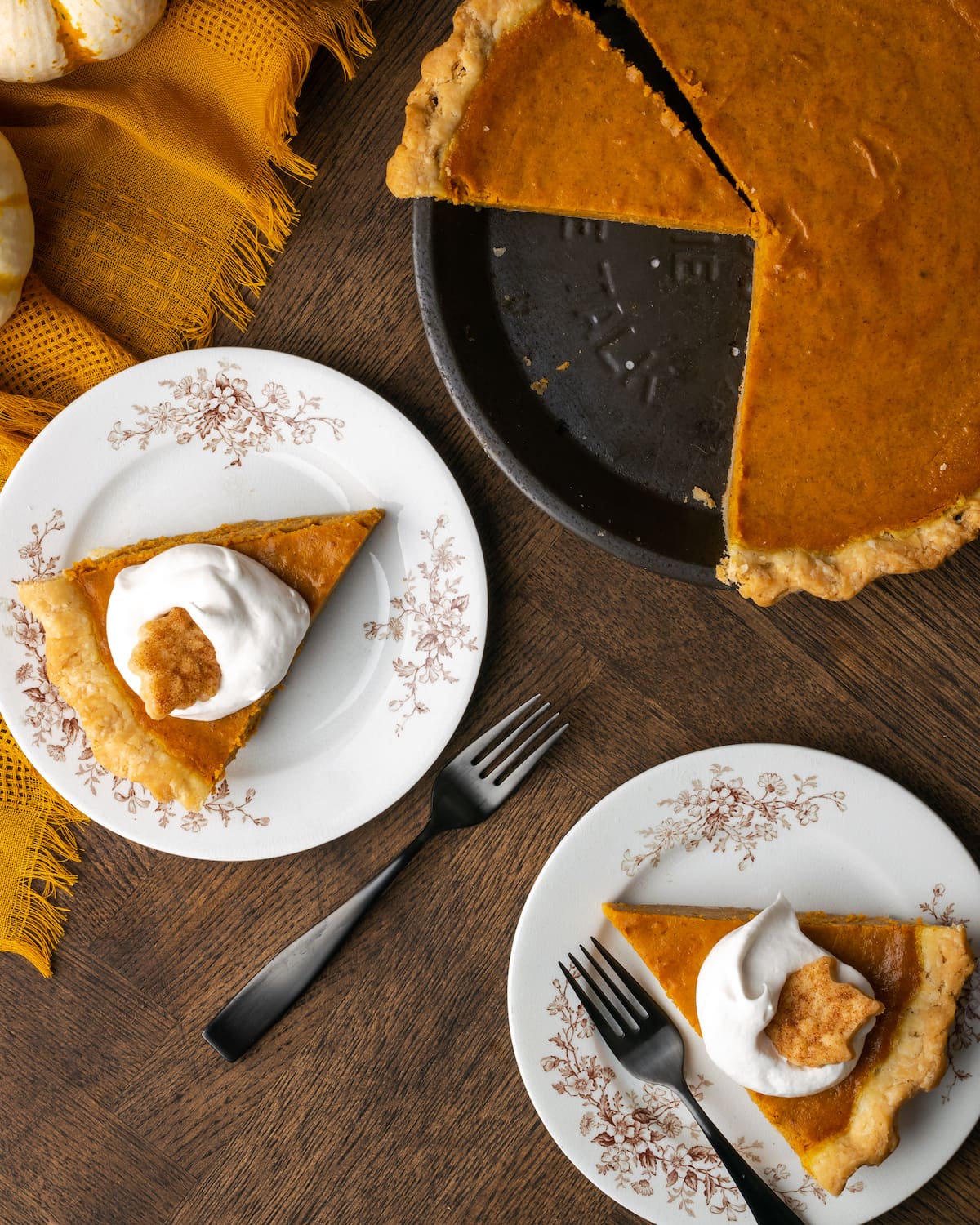 Vista aérea de dos rebanadas de pastel de calabaza en platos, cubiertos con crema batida, junto a un pastel de calabaza al que le faltan rebanadas.