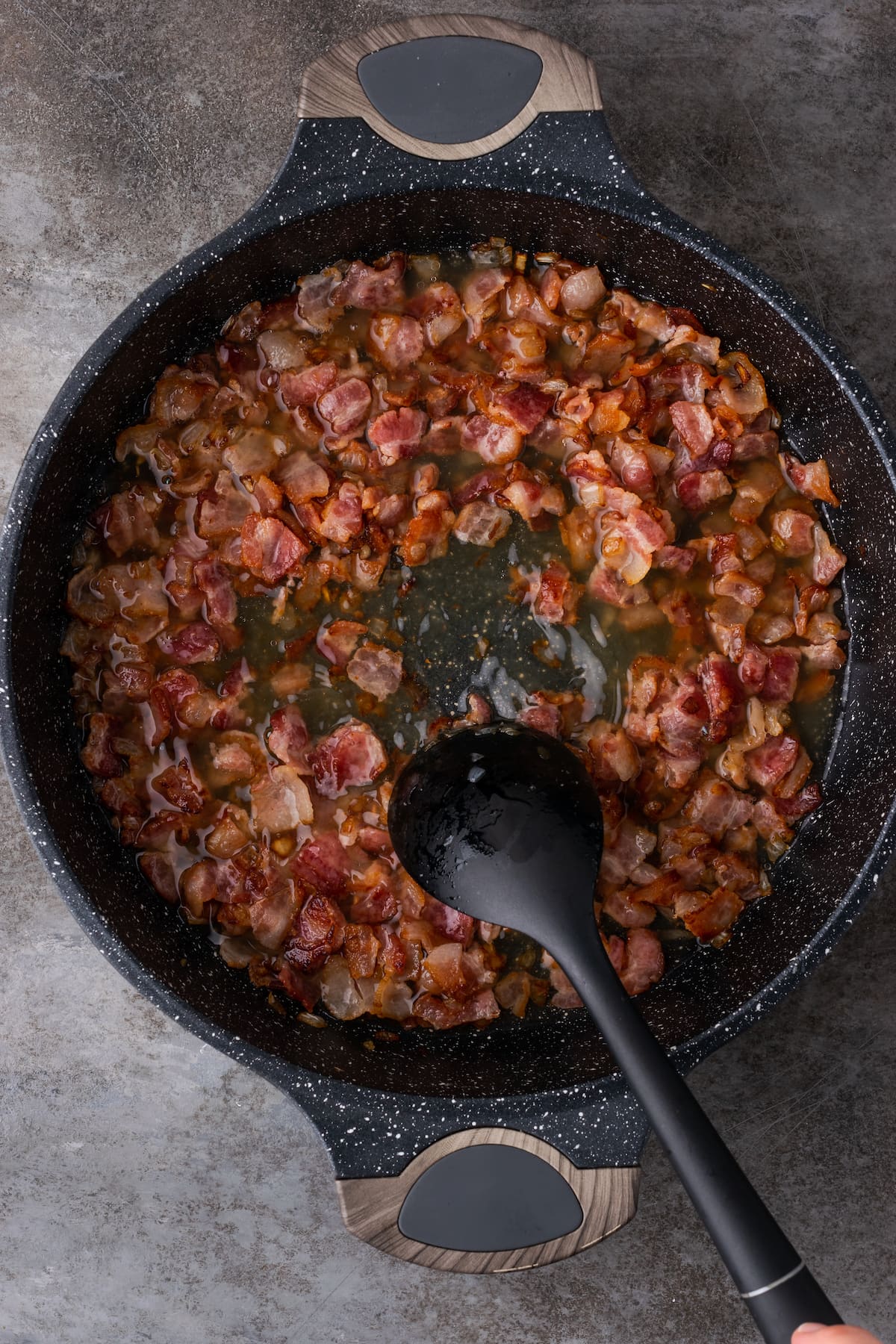 Una cuchara de plástico raspa el fondo de una sartén llena de cebolla caramelizada y tocino, glaseada con caldo y vino blanco.