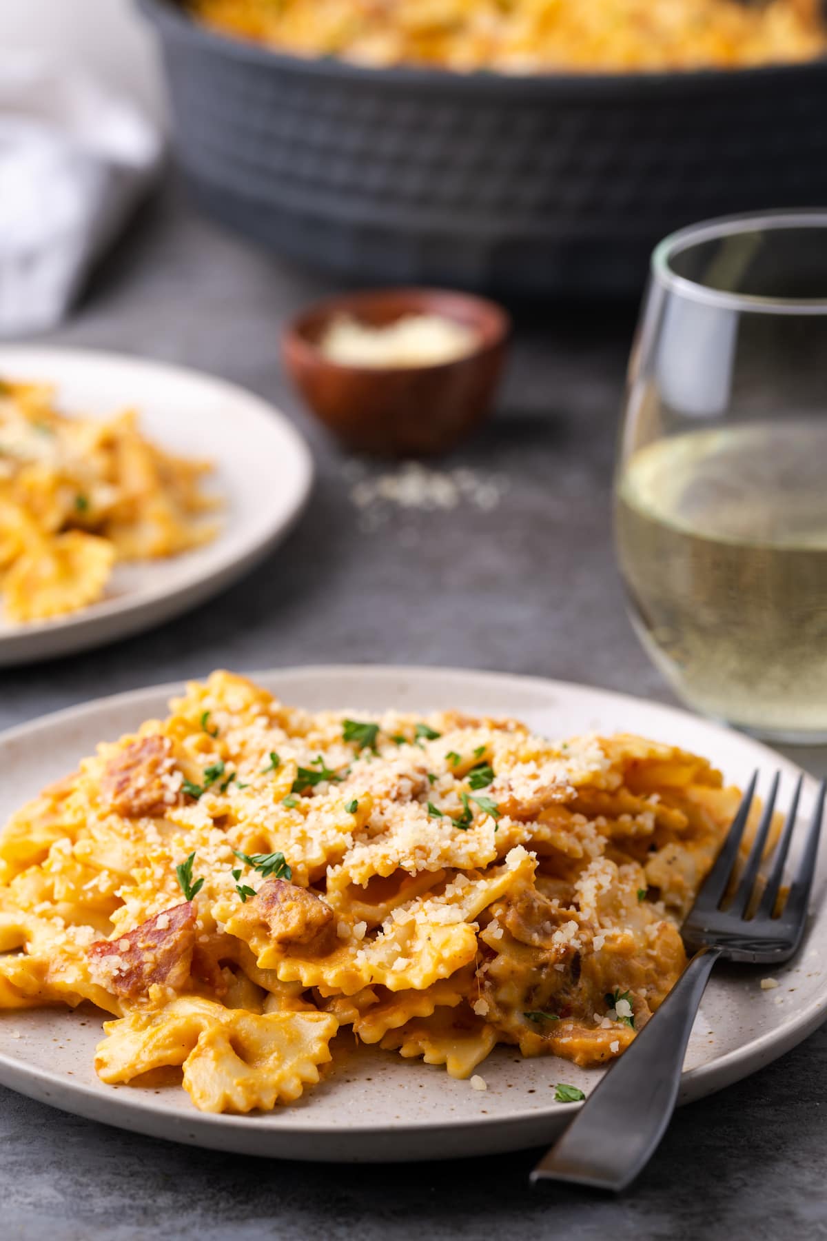 Una porción de pasta de calabaza con tocino en un plato junto a un tenedor, con una copa de vino blanco de fondo.