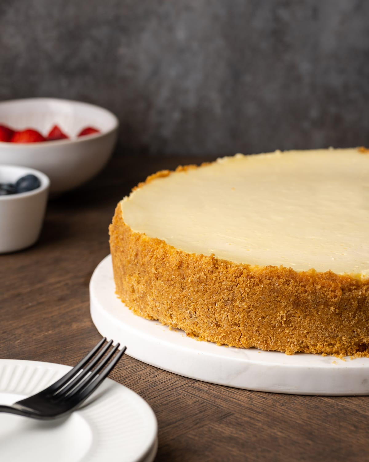 Un pastel de queso con crema agria entero en un plato blanco, al lado de un plato con un tenedor.