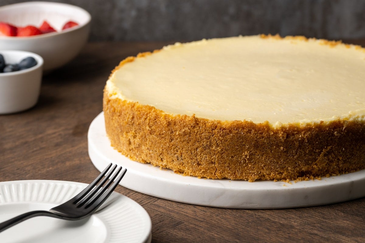 Un pastel de queso con crema agria entero en un plato blanco, al lado de un plato con un tenedor.