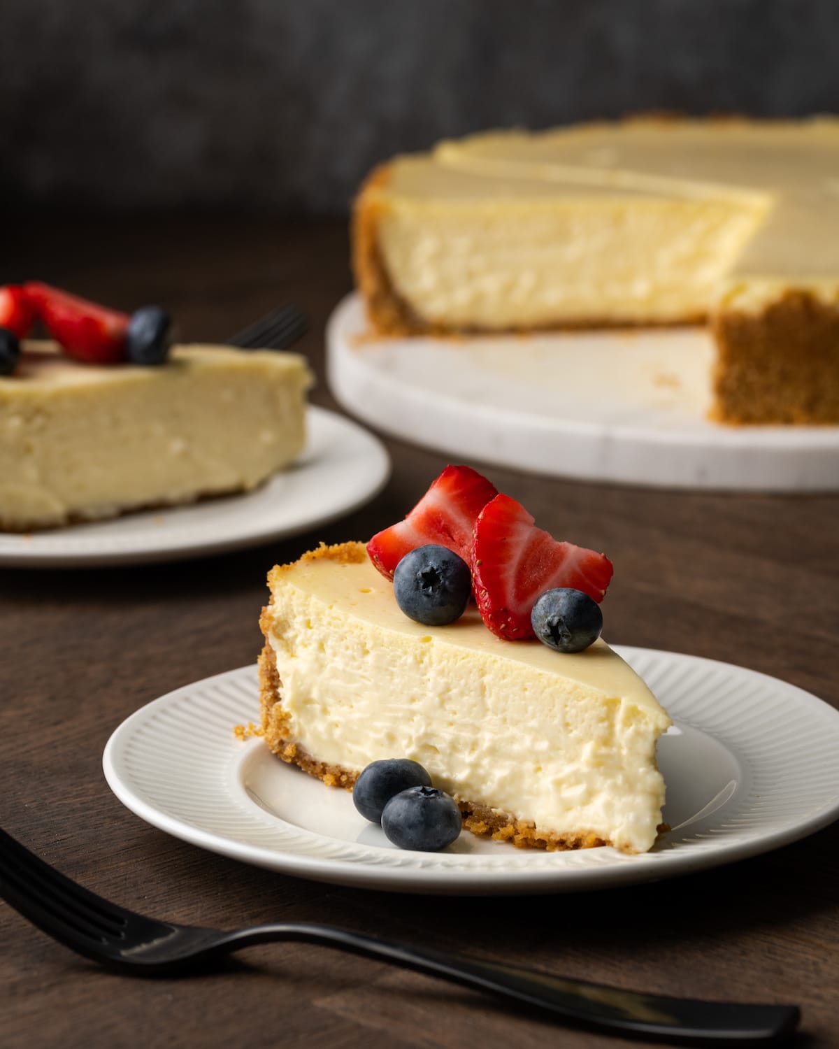 Una rebanada de tarta de queso con crema agria adornada con bayas frescas en un plato blanco, con la tarta de queso completa al fondo.