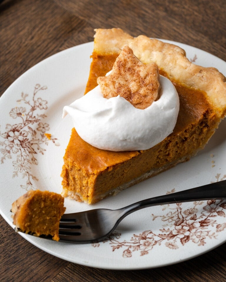 Un trozo de pastel de calabaza en un plato cubierto con crema batida y una galleta de azúcar, al que le falta un tenedor lleno de pastel.