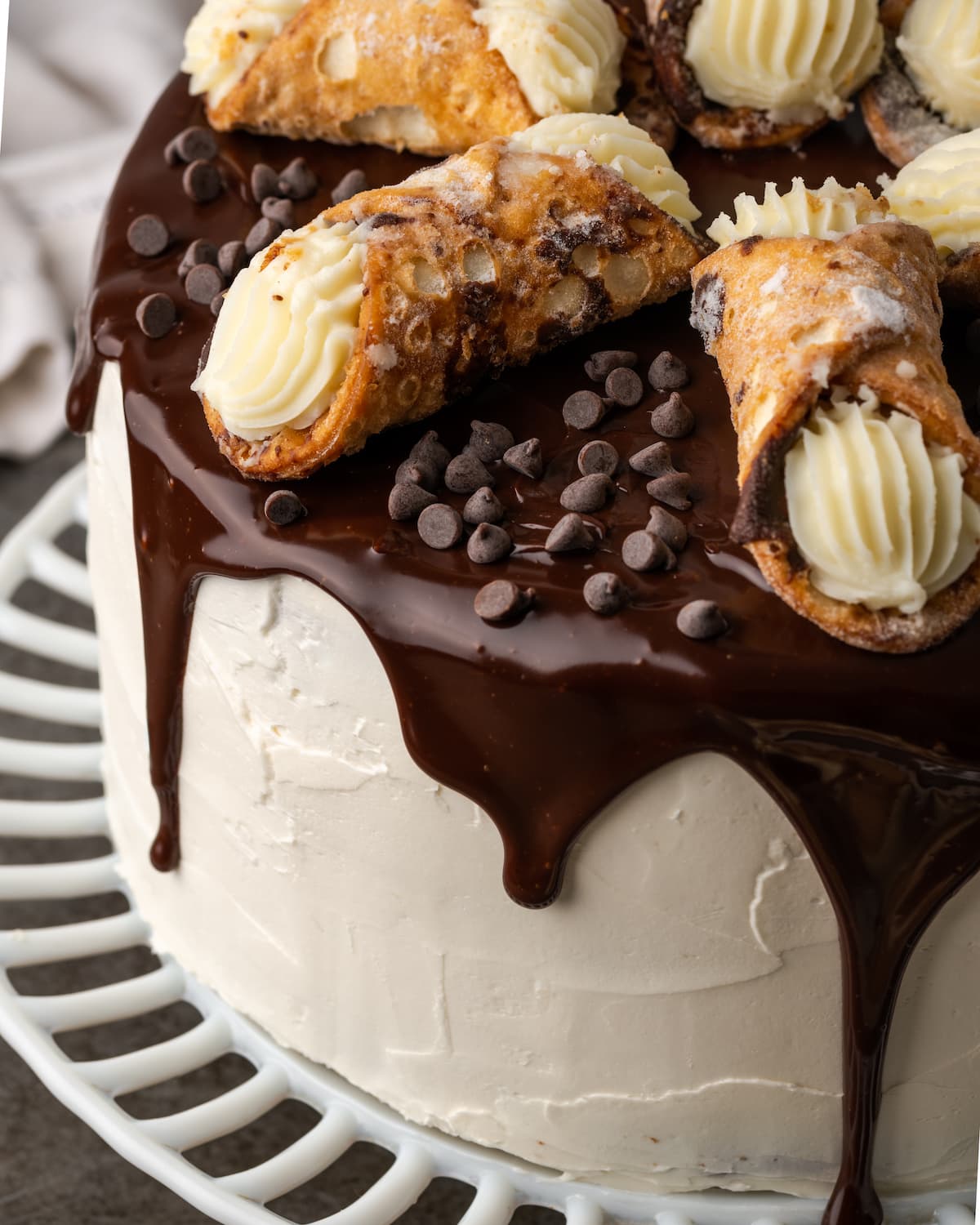 Esta receta de pastel Cannoli transforma un favorito de la panadería en un delicioso pastel de capas de vainilla. ¡Completa con relleno de mascarpone casero! Vista superior del pastel de cannoli en un puesto de pasteles rociado con ganache de chocolate y adornado con pasteles de cannoli.