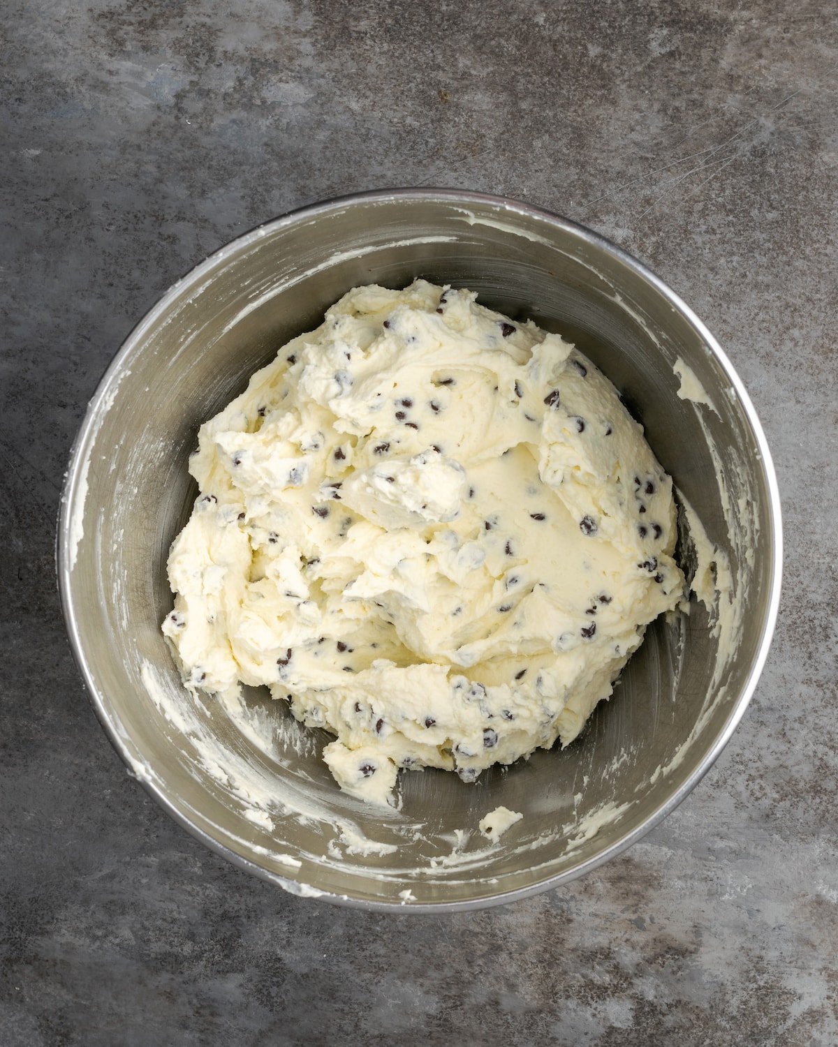 Esta receta de pastel Cannoli transforma un favorito de la panadería en un delicioso pastel de capas de vainilla. ¡Completa con relleno de mascarpone casero! Chispas de chocolate agregadas a los ingredientes de relleno de mascarpone en un tazón de metal.