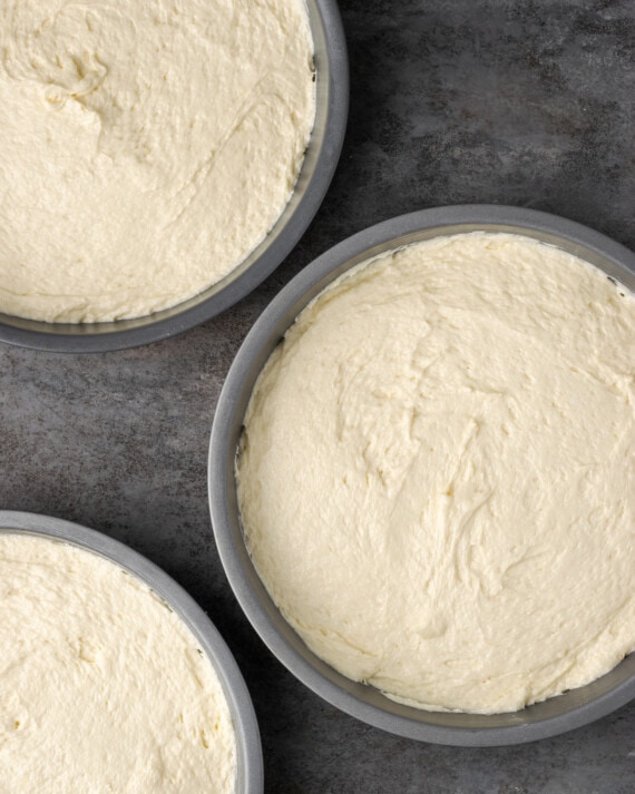 Esta receta de pastel Cannoli transforma un favorito de la panadería en un delicioso pastel de capas de vainilla. ¡Completa con relleno de mascarpone casero! Masa para pastel de cannoli dividida entre tres moldes redondos para pastel.