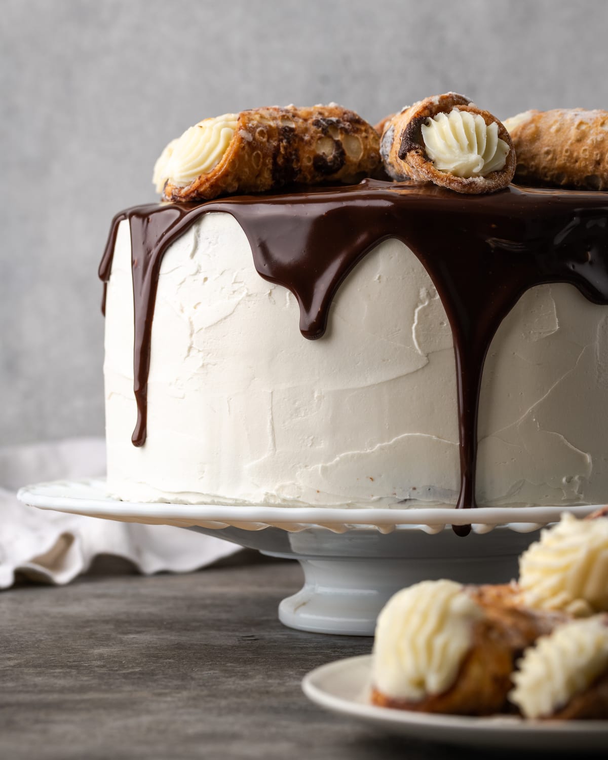 Esta receta de pastel Cannoli transforma un favorito de la panadería en un delicioso pastel de capas de vainilla. ¡Completa con relleno de mascarpone casero! Pastel de cannoli en un soporte para pasteles rociado con ganache de chocolate y adornado con pasteles de cannoli.