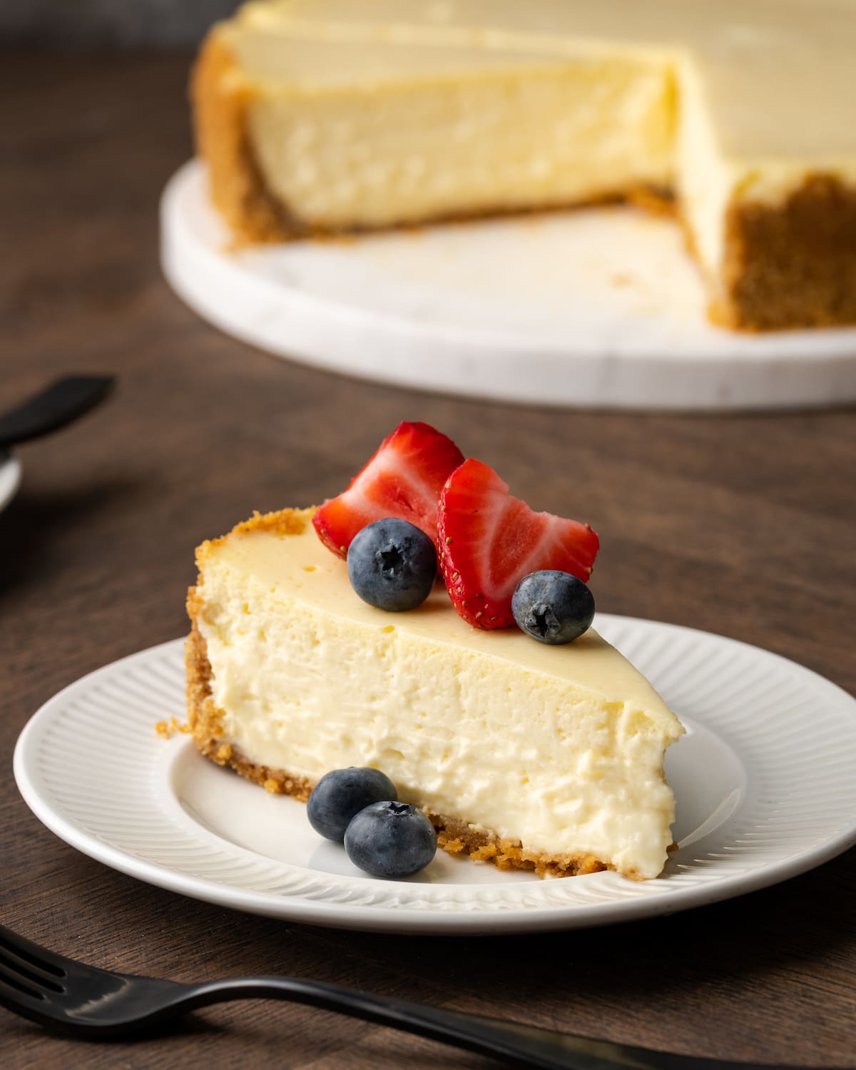 Una rebanada de tarta de queso con crema agria adornada con bayas frescas en un plato blanco, con la tarta de queso completa al fondo.