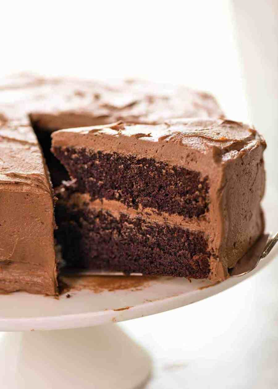 Una foto que muestra la mitad de un pastel de chocolate glaseado con crema de mantequilla de chocolate en un soporte de pastel blanco con una porción grande parcialmente extraída.