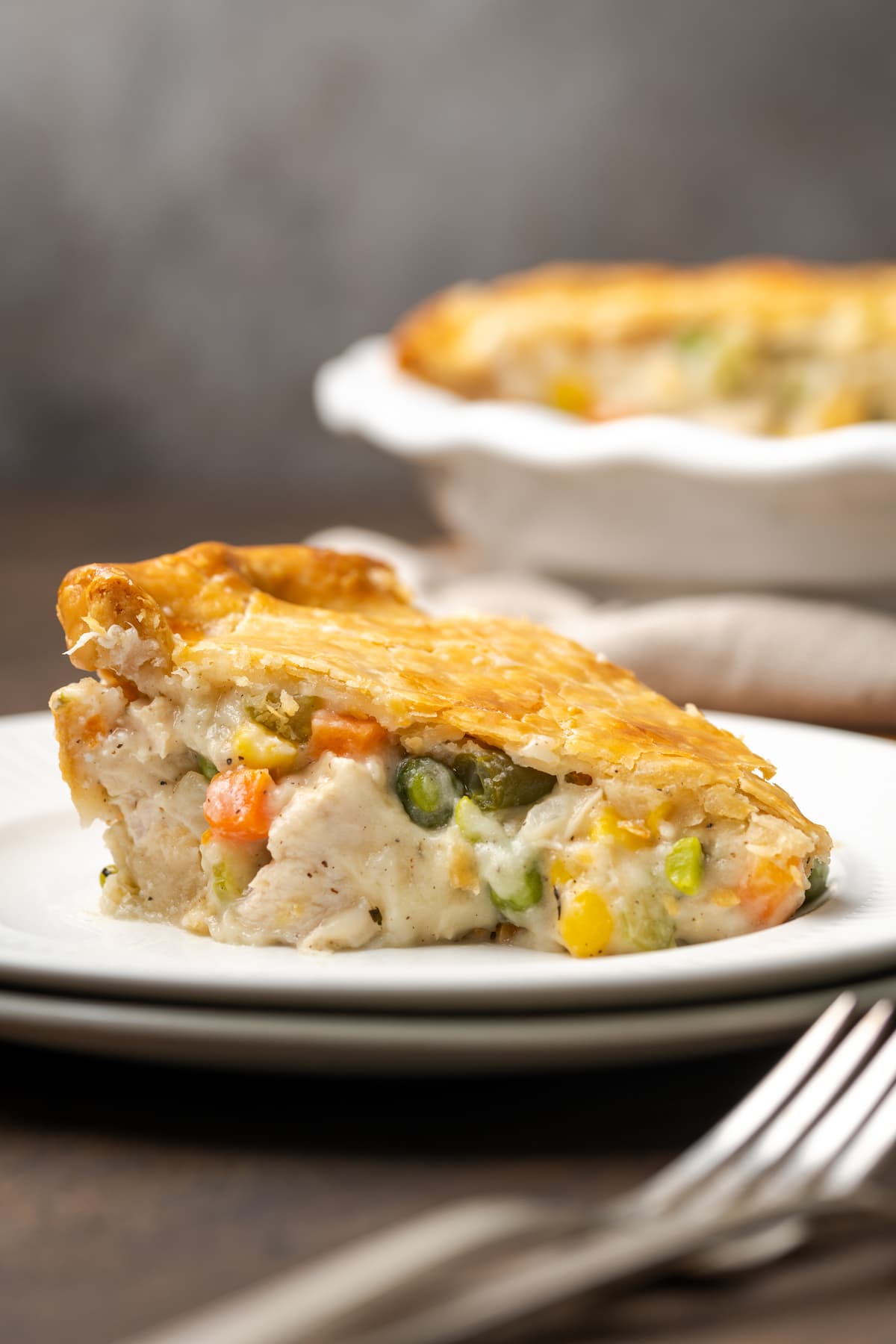 El Chicken Pot Pie casero es la mejor comida reconfortante, hecho con pollo cremoso y verduras horneadas dentro de una masa de pastel de hojaldre mantecosa. Un trozo de pastel de pollo en un plato blanco junto a un tenedor.