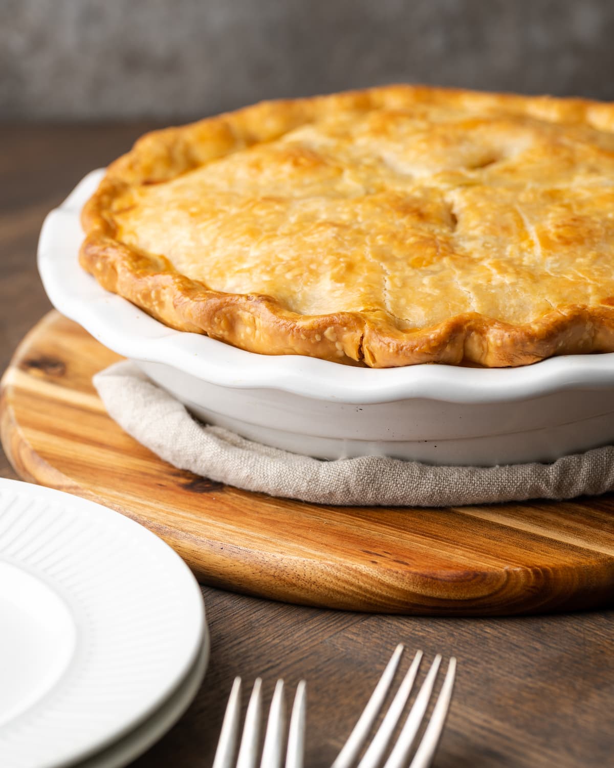 El Chicken Pot Pie casero es la mejor comida reconfortante, hecho con pollo cremoso y verduras horneadas dentro de una masa de pastel de hojaldre mantecosa. Pastel de pollo al horno dentro de un molde para pastel junto a platos blancos apilados y dos tenedores.