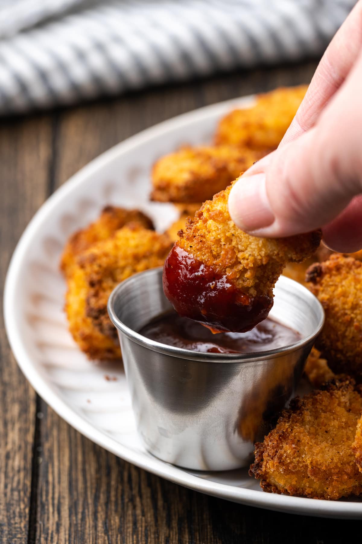 Crispy Air Fryer Popcorn Chicken es irresistiblemente crujiente por fuera y jugoso por dentro, ¡listo en 20 minutos de principio a fin! Una mano que sostiene un trozo de pollo con palomitas de maíz parcialmente sumergido en un molde de salsa BBQ, en un plato con más trozos de pollo con palomitas de maíz.