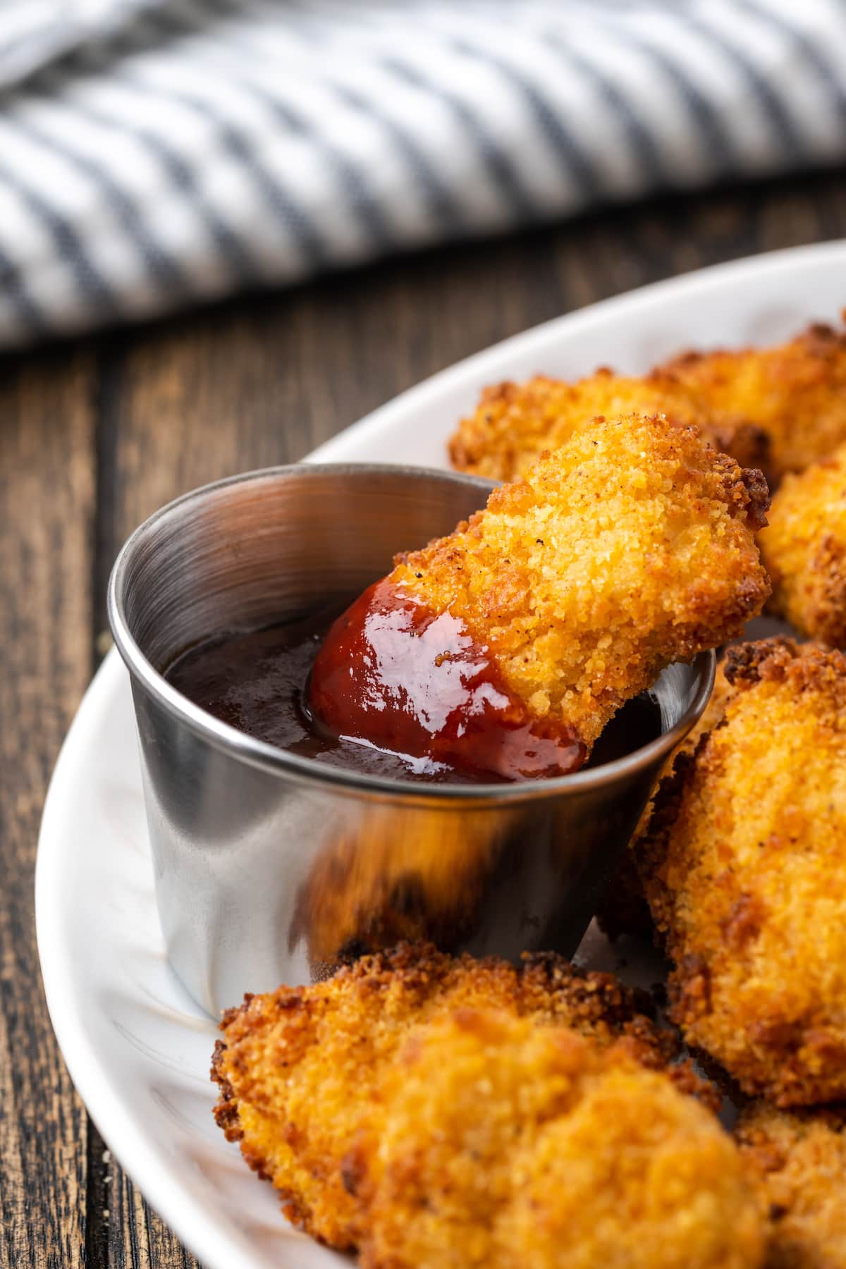 Crispy Air Fryer Popcorn Chicken es irresistiblemente crujiente por fuera y jugoso por dentro, ¡listo en 20 minutos de principio a fin! Un trozo de pollo con palomitas de maíz parcialmente sumergido en un molde de salsa BBQ, en un plato con más trozos de pollo con palomitas de maíz.