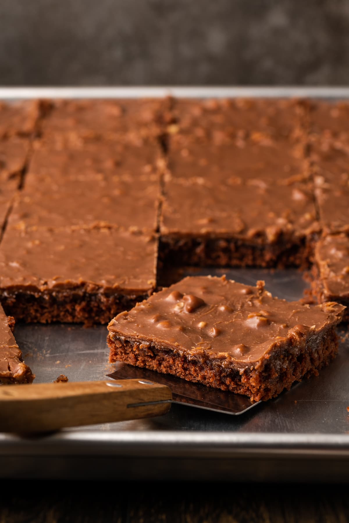 Fudgy Texas Sheet Cake es el sueño de los amantes de los pasteles de chocolate. Esta receta fácil de pastel está cubierta con glaseado de chocolate y es perfecta para fiestas. Una rebanada de pastel de hoja de Texas se levanta de una bandeja con una espátula.