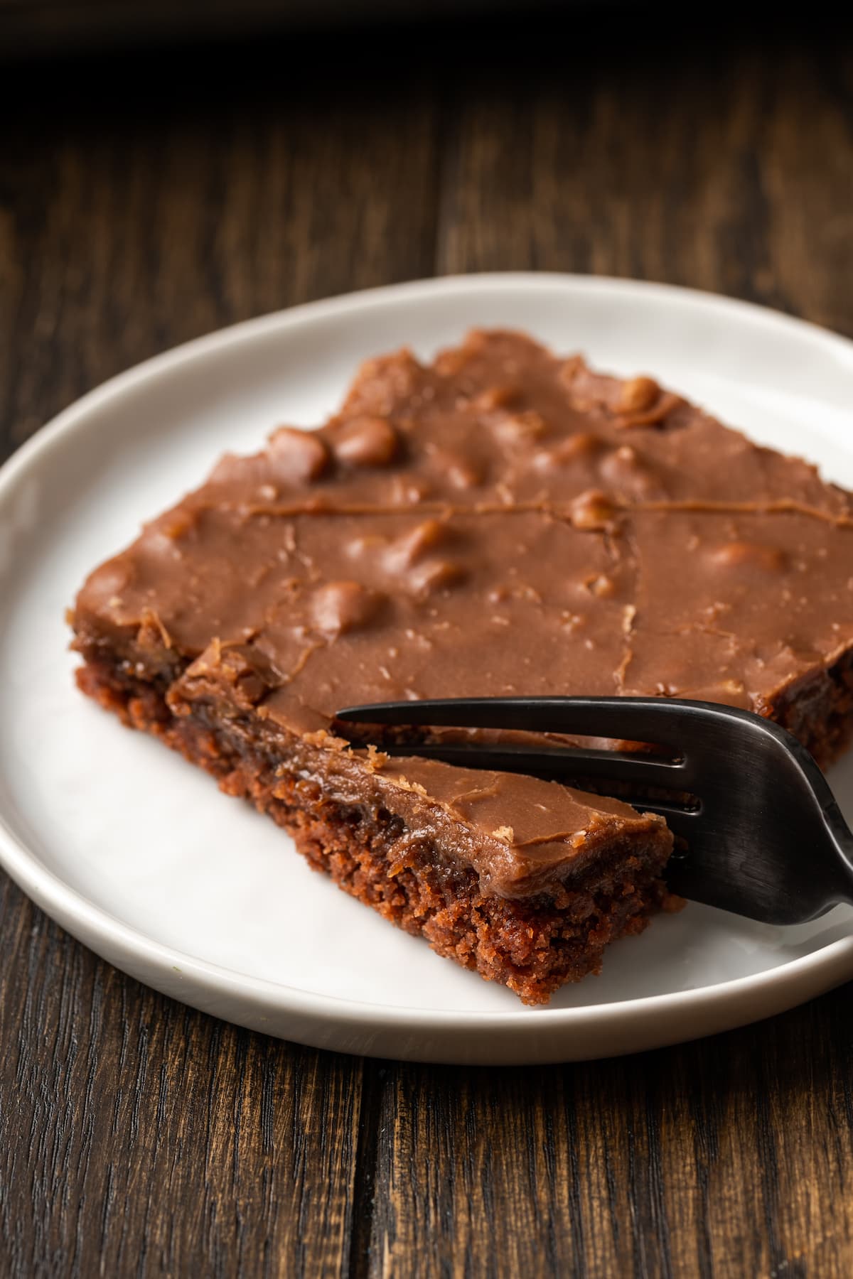 Fudgy Texas Sheet Cake es el sueño de los amantes de los pasteles de chocolate. Esta receta fácil de pastel está cubierta con glaseado de chocolate y es perfecta para fiestas. Un tenedor se clava en la esquina de una rebanada de pastel de hojas de Texas en un plato blanco.