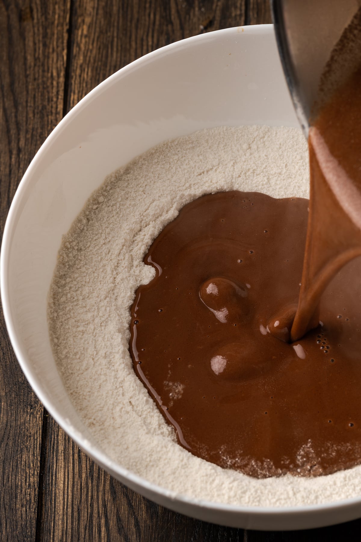 Fudgy Texas Sheet Cake es el sueño de los amantes de los pasteles de chocolate. Esta receta fácil de pastel está cubierta con glaseado de chocolate y es perfecta para fiestas. La mantequilla caliente mezclada con cacao se vierte en un tazón de ingredientes secos para la masa para pasteles.