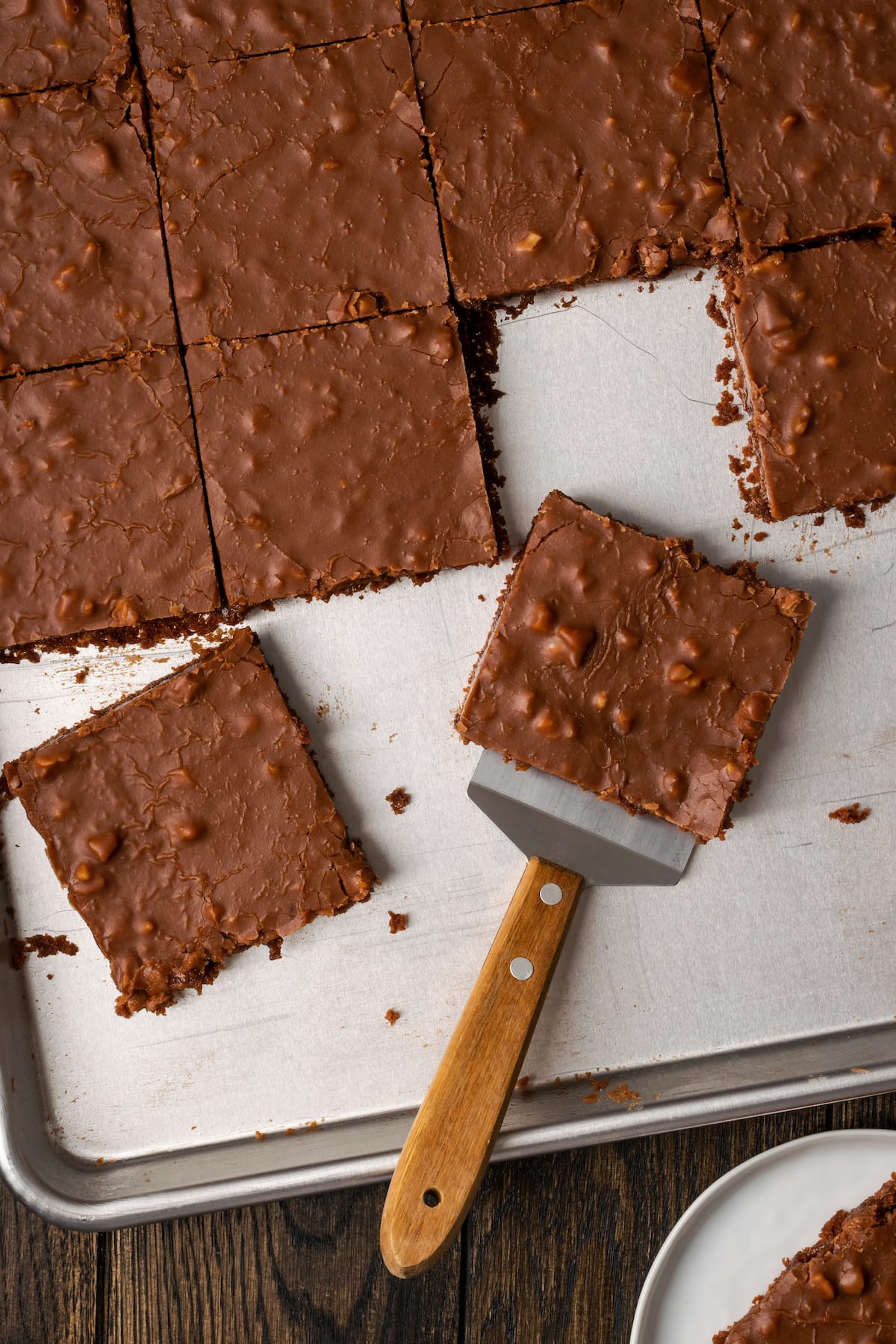 Fudgy Texas Sheet Cake es el sueño de los amantes de los pasteles de chocolate. Esta receta fácil de pastel está cubierta con glaseado de chocolate y es perfecta para fiestas. Vista aérea de una rebanada de pastel de hoja de Texas que se sirve de una bandeja con una espátula.