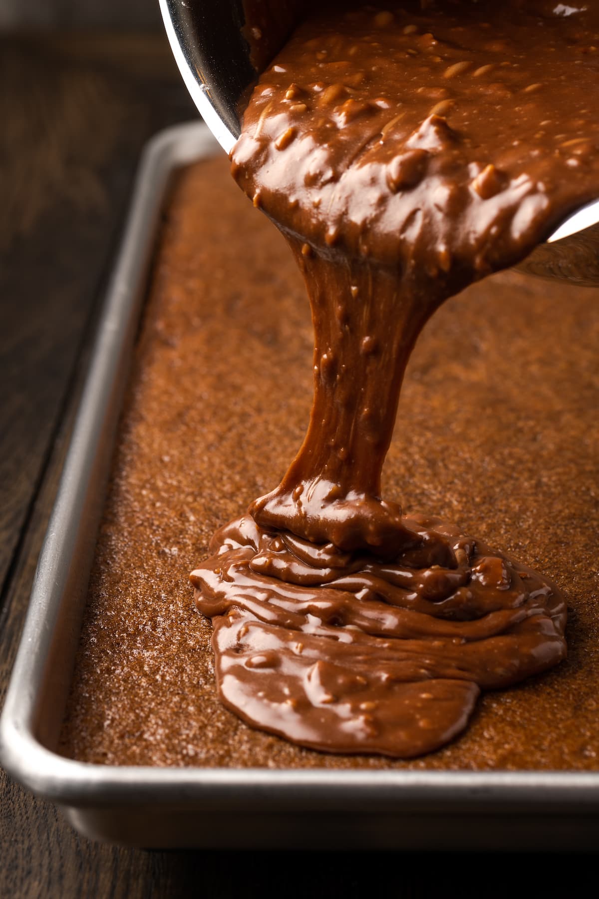 Fudgy Texas Sheet Cake es el sueño de los amantes de los pasteles de chocolate. Esta receta fácil de pastel está cubierta con glaseado de chocolate y es perfecta para fiestas. El glaseado de chocolate con nueces se vierte sobre un pastel de hojas de Texas recién horneado.