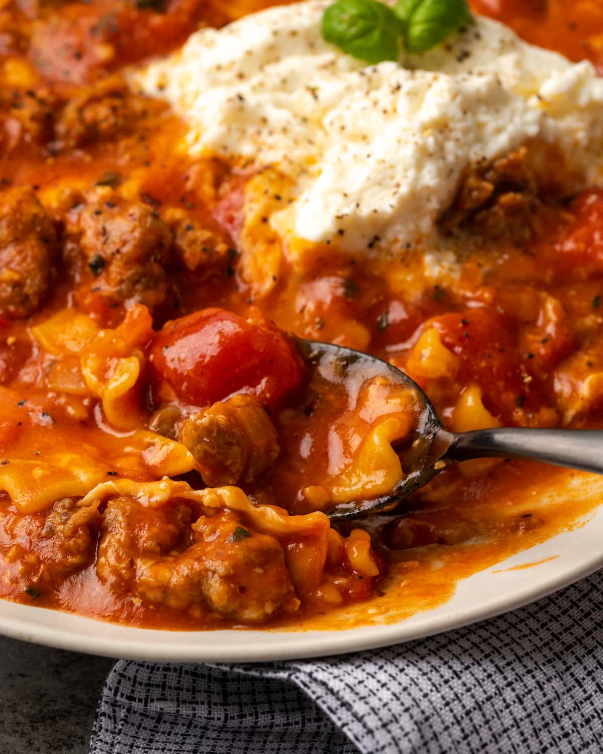 La sopa de lasaña es todo lo que amamos de lasaña casera envuelta en una comida de una sola olla. ¡Prepara esta receta fácil de sopa en 30 minutos o menos! Un tazón de sopa de lasaña cubierto con queso ricotta y hojas de albahaca, con una cuchara.