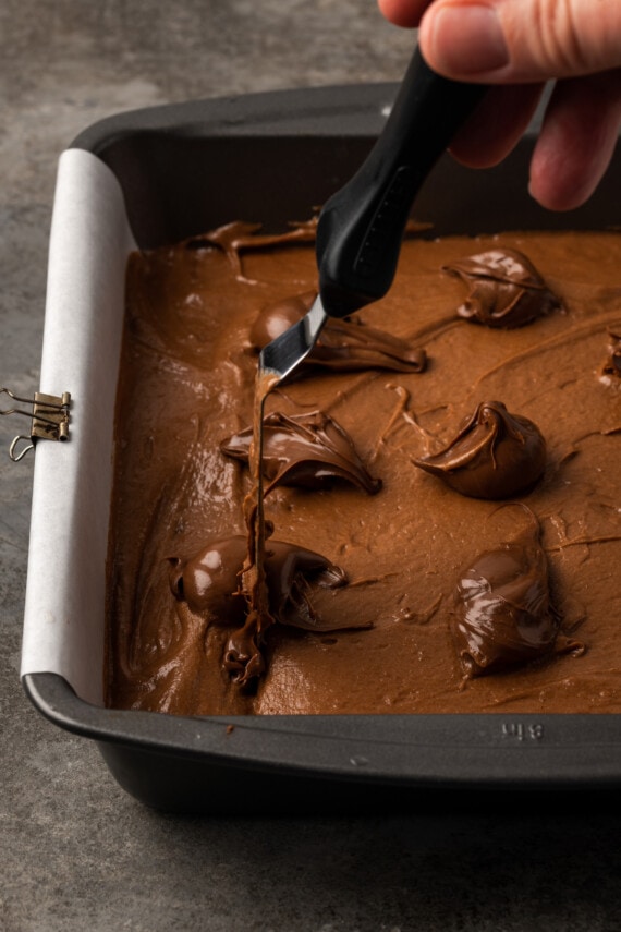 Los brownies caseros de Nutella son ricos, dulces y se arremolinan con crema de chocolate y avellanas. ¡Prepáralos en un tazón con solo 7 ingredientes! Se usa una espátula desplazada para hacer girar cucharadas de Nutella en una sartén llena de masa de brownie.