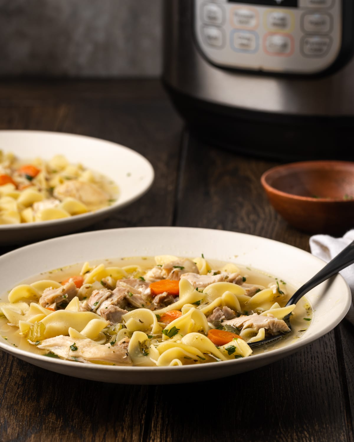 Instant Pot Chicken Noodle Soup es un atajo rápido y fácil para un alimento reconfortante favorito. Rebosante de ingredientes clásicos y toneladas de sabor. Un tazón de sopa de fideos con pollo Instant Pot, con un segundo tazón y el Instant Pot en el fondo.