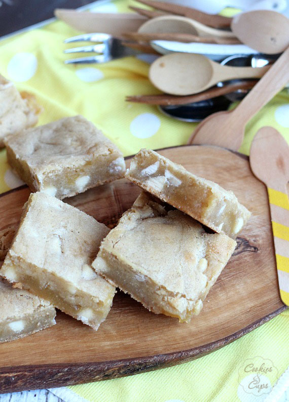 Blondies surtidos de chocolate blanco y limón dispuestos en una fuente de madera para servir, con utensilios en el fondo.