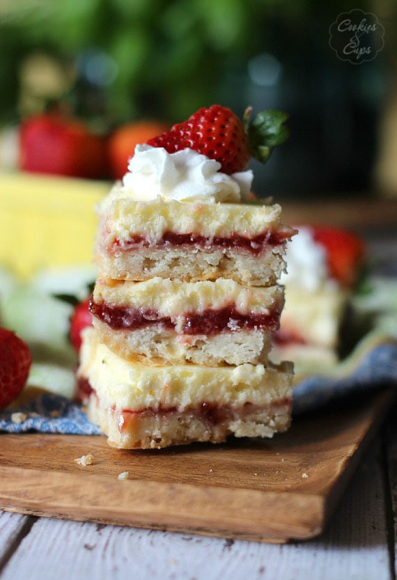 Una pila de tres barras de tarta de queso con limón y fresa cubiertas con crema batida y una fresa en una tabla de madera.