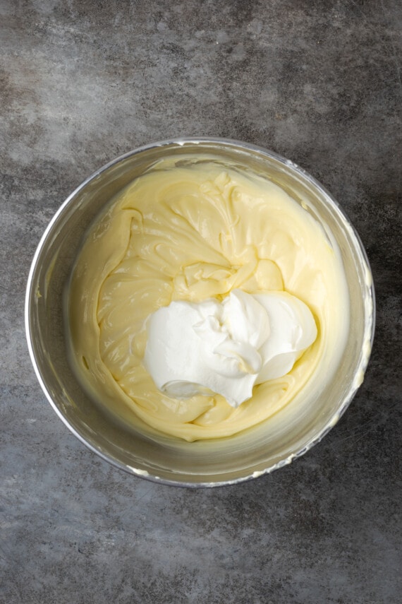 Banana Blondie Cheesecake es un postre fácil con cheesecake de vainilla al horno empapado en caramelo salado, en capas sobre una gruesa banana blondie. Crema agria agregada a la masa de tarta de queso en un tazón.