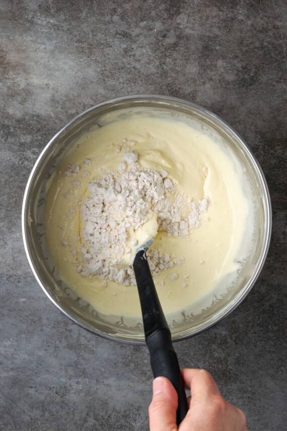 Banana Blondie Cheesecake es un postre fácil con cheesecake de vainilla al horno empapado en caramelo salado, en capas sobre una gruesa banana blondie. La harina se revuelve en la masa de tarta de queso en un tazón para mezclar.