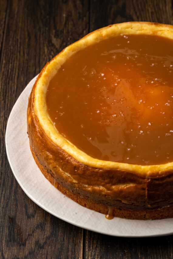 Banana Blondie Cheesecake es un postre fácil con cheesecake de vainilla al horno empapado en caramelo salado, en capas sobre una gruesa banana blondie. El cheesecake de banana blondie ensamblado en un soporte para pasteles.