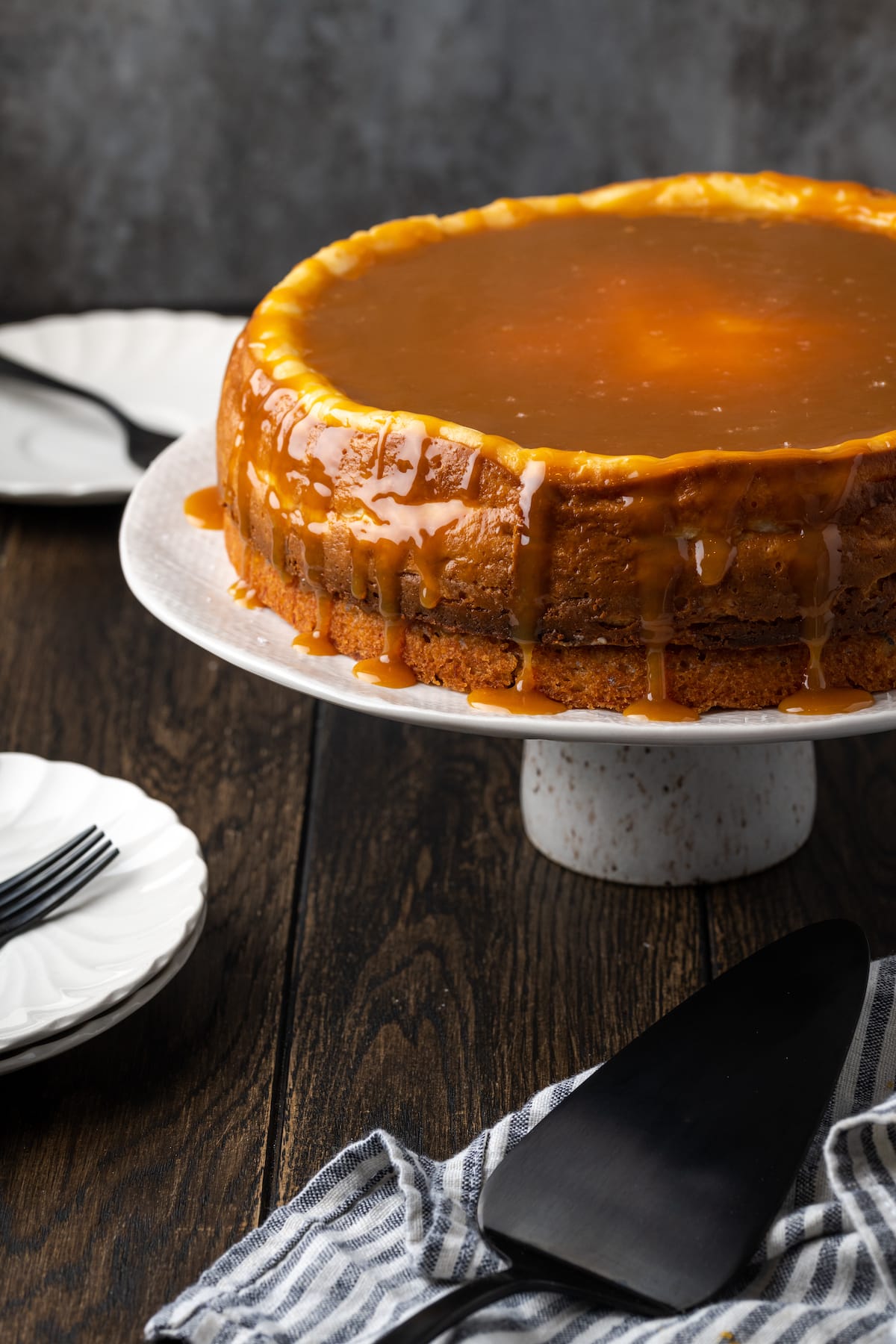 Banana Blondie Cheesecake es un postre fácil con cheesecake de vainilla al horno empapado en caramelo salado, en capas sobre una gruesa banana blondie. Cheesecake de banana blondie cubierto con caramelo en un soporte de pastel blanco, colocado encima de una encimera de madera oscura al lado de un cuchillo para pastel.