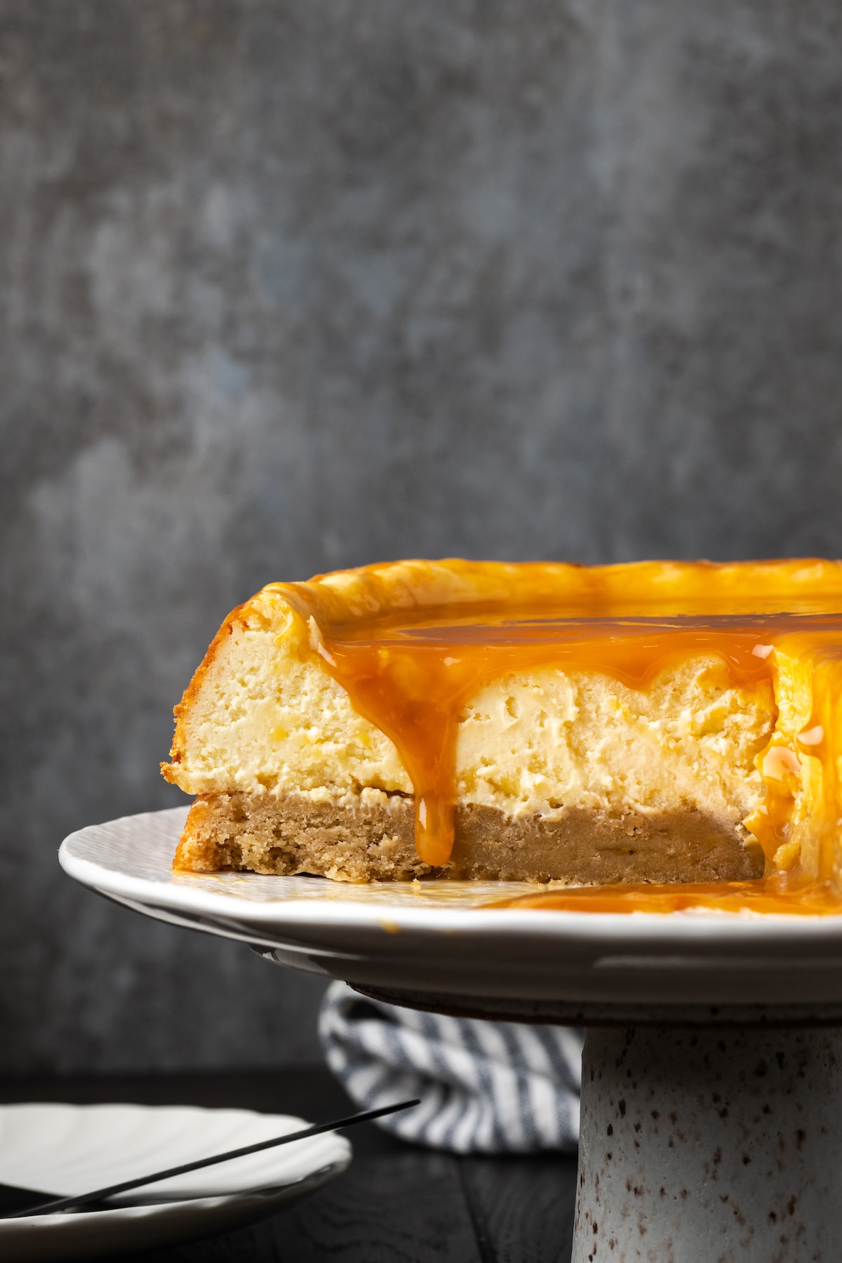 Banana Blondie Cheesecake es un postre fácil con cheesecake de vainilla al horno empapado en caramelo salado, en capas sobre una gruesa banana blondie. Cheesecake de banana blondie cubierto con caramelo en un soporte para pasteles, al que le falta una rebanada.