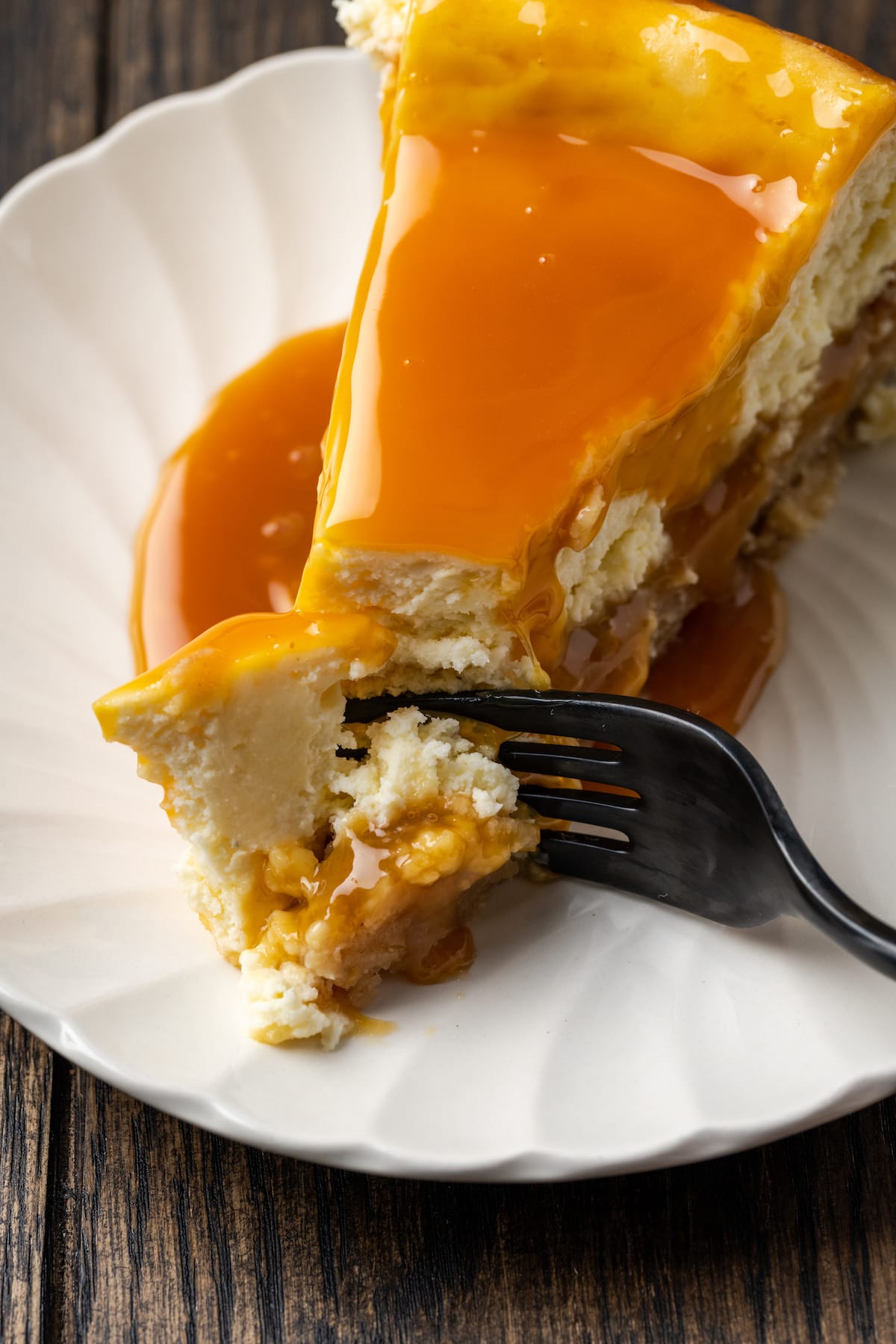 Banana Blondie Cheesecake es un postre fácil con cheesecake de vainilla al horno empapado en caramelo salado, en capas sobre una gruesa banana blondie. Un tenedor corta la punta de una rebanada de pastel de queso rubio con plátano empapado en caramelo en un plato blanco.