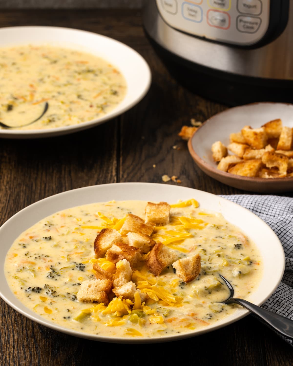 Un tazón de sopa instantánea de brócoli y queso cheddar cubierto con picatostes con una cuchara, con un segundo tazón de sopa junto a un tazón de picatostes en el fondo.