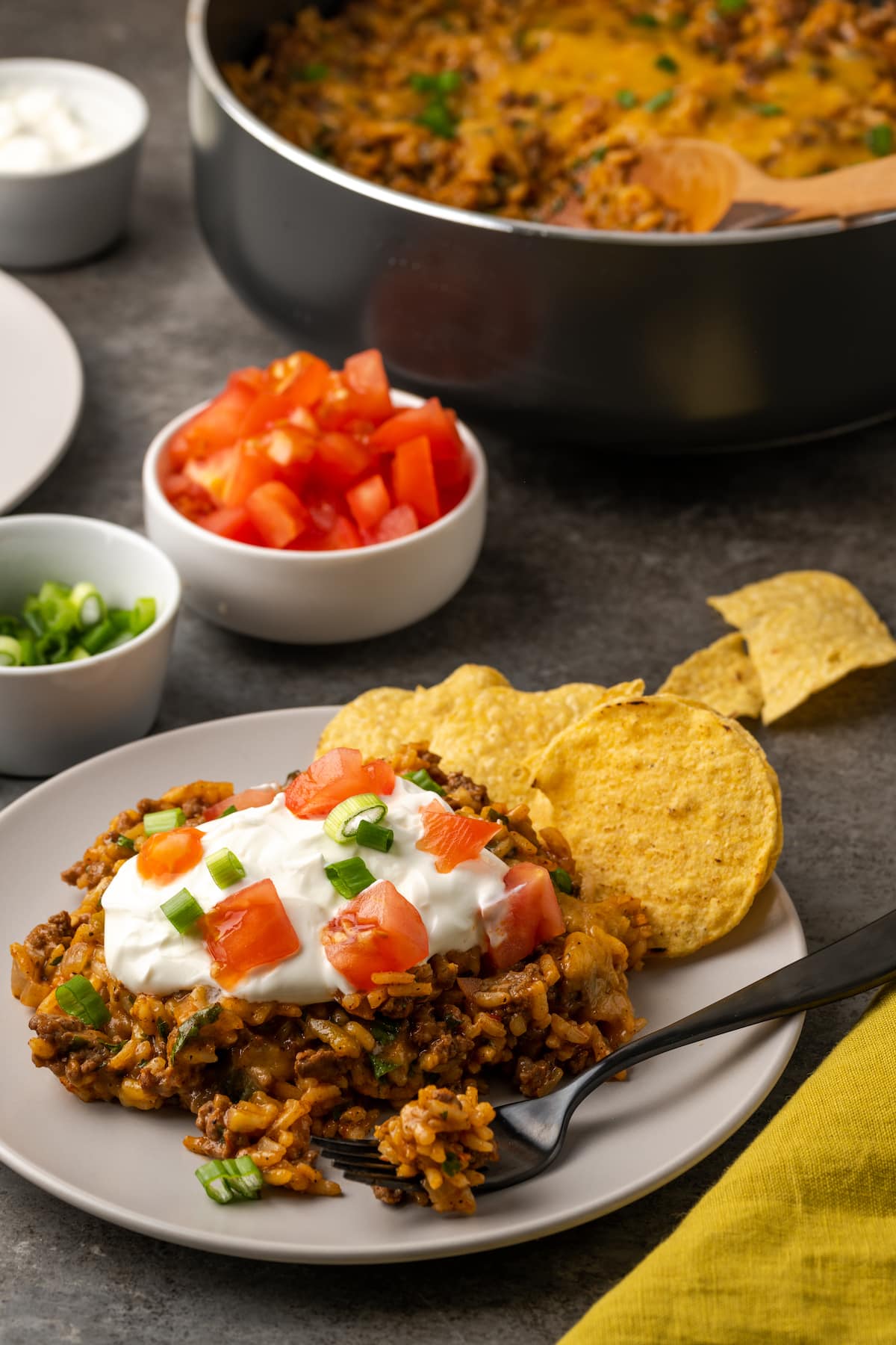 Una porción de taco de arroz con queso en un plato junto a chips de maíz, cubierto con crema agria, tomates cortados en cubitos y cebollas verdes con tazones de ingredientes y una sartén en el fondo.
