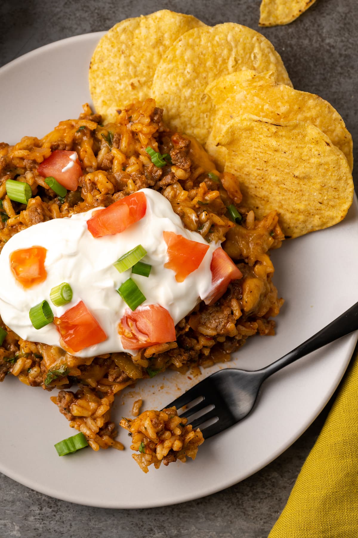 Vista aérea de tacos de arroz con queso en un plato cubierto con crema agria, tomates cortados en cubitos y cebollas verdes junto a chips de maíz y un tenedor.