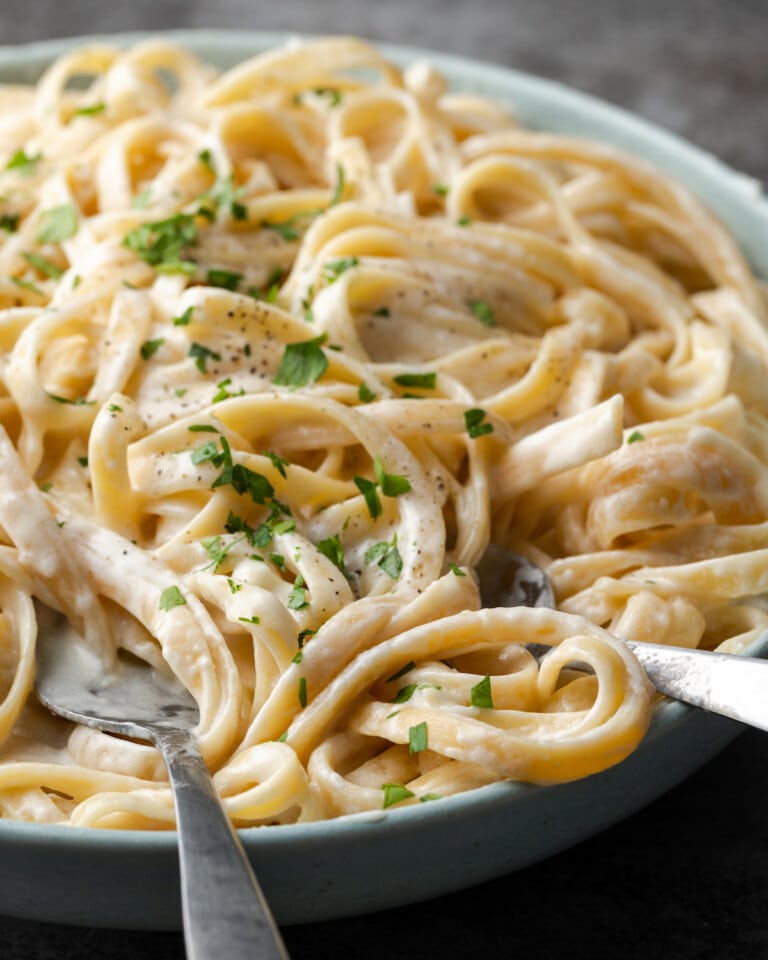 Un tazón de fettuccine Alfredo adornado con perejil fresco picado, con utensilios de plata para servir.