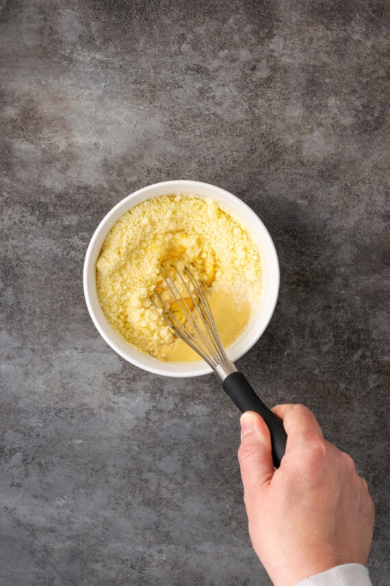 Una mano usando un batidor para combinar los ingredientes de la salsa de pasta César en un tazón.